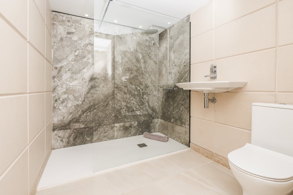 Modern bathroom featuring a walk-in shower with a glass partition and grey marble-effect wall. Includes minimalist white fixtures and cream-toned tiling.