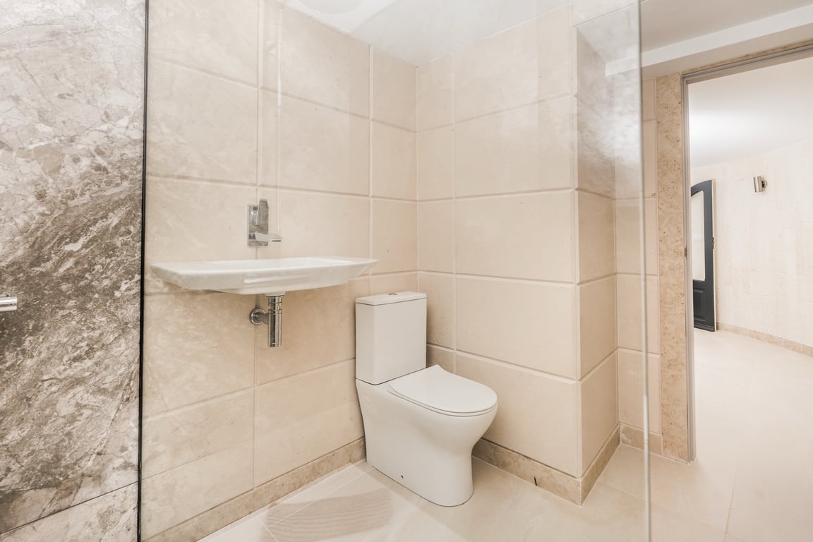 Modern bathroom featuring cream limestone-effect tiling, a contemporary floor-mounted toilet, and a wall-hung sink. Includes a glass shower partition and marble accents.