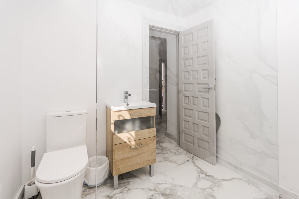 Modern bathroom featuring white marble-effect tiling, a light wood vanity unit, and a frameless glass shower partition for a clean, bright finish.