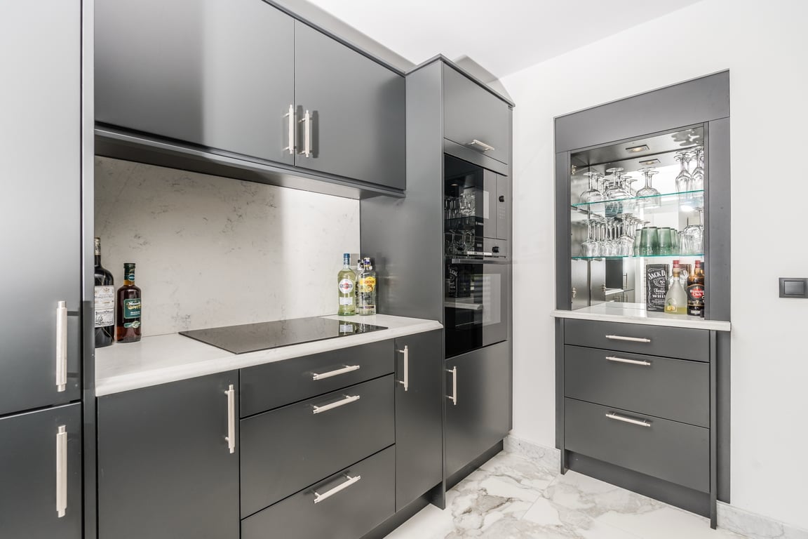 Modern kitchen featuring charcoal grey cabinetry, white marble countertops, and integrated appliances. A built-in bar area with a glass display cabinet complements the marble flooring.