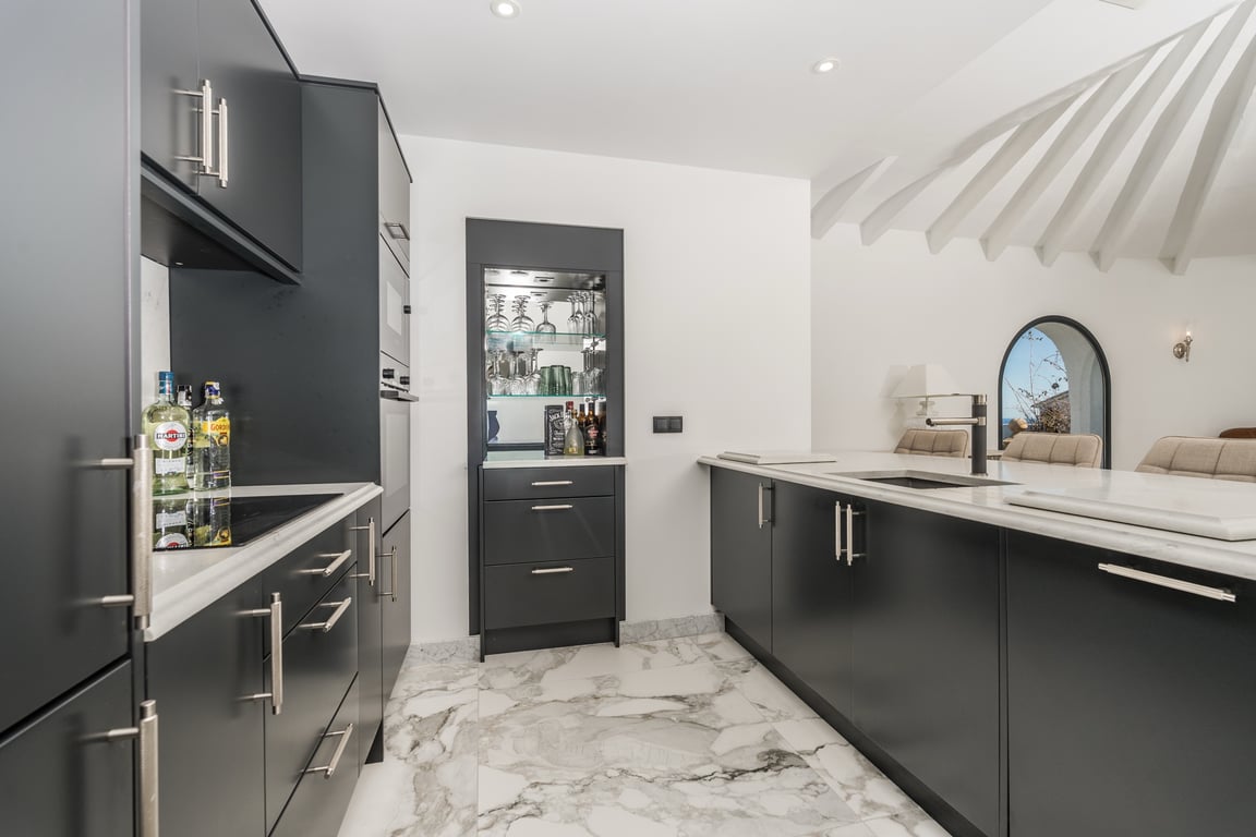 Modern kitchen with dark matte cabinetry, marble surfaces, and a central island. Features include an integrated bar niche and recessed lighting against a radial beamed ceiling.