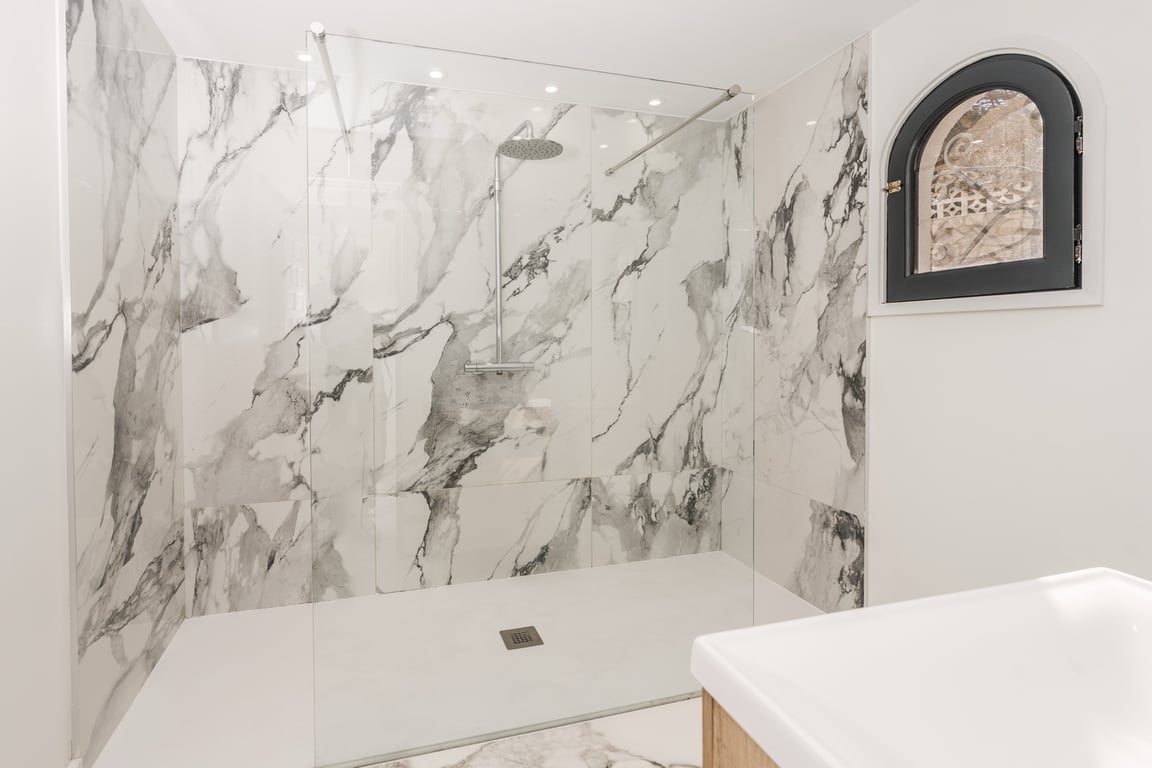 Modern walk-in shower featuring marble-veined wall tiles, a sleek glass partition, and a traditional arched window with a black frame and wrought iron detail.