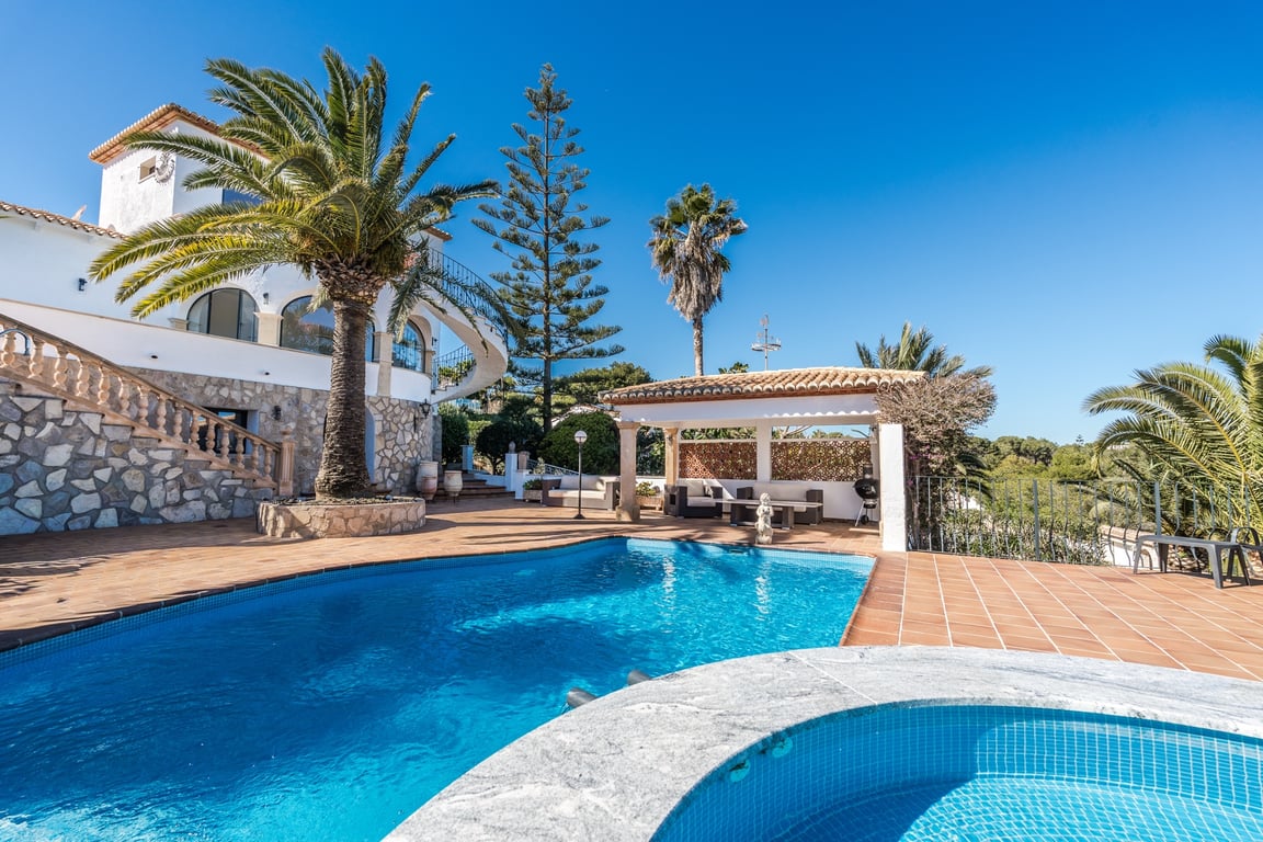 Outdoor pool area featuring a built-in jacuzzi, terracotta terrace, and Mediterranean garden with palms, set against a stone-clad villa with a covered lounge.