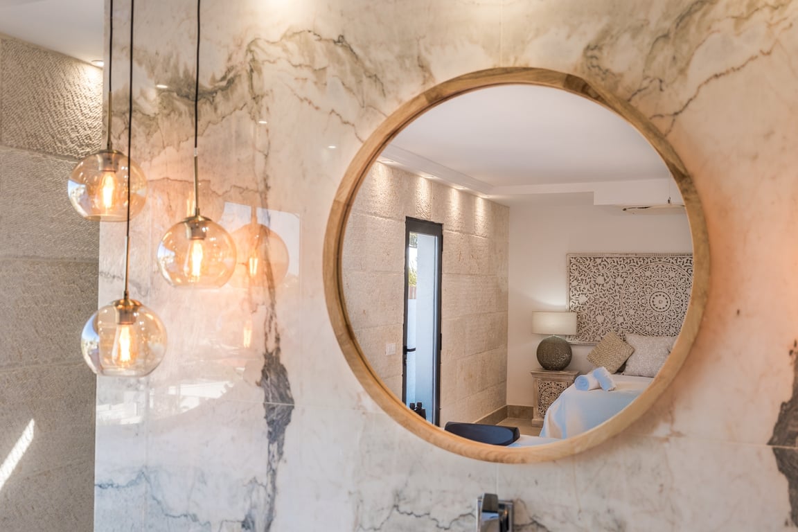 Round wood-framed mirror mounted on a veined marble wall, featuring glass globe pendant lights and a reflection of the bedroom with textured limestone.