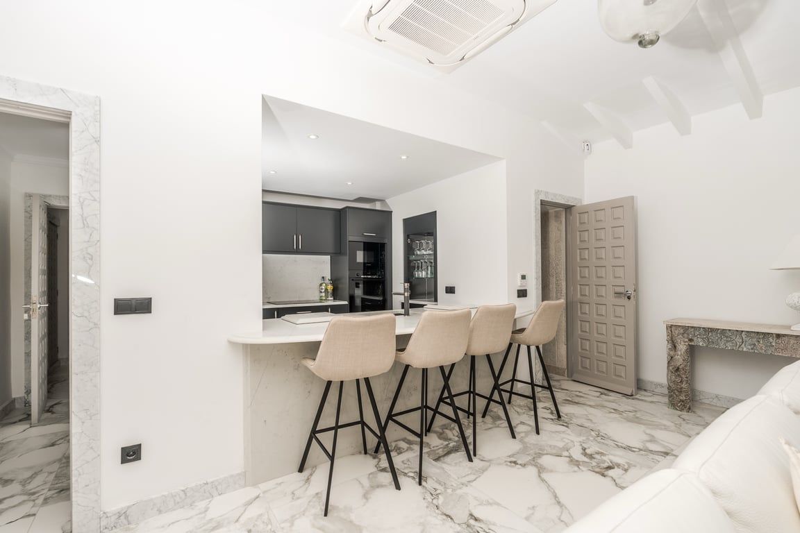 Semi-open kitchen featuring a marble breakfast bar with four stools, dark cabinetry, and integrated lighting. Finished with marble flooring and white-beamed ceilings.