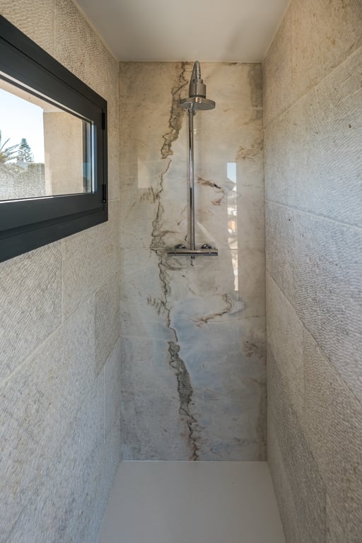 Walk-in shower featuring a marble accent wall with prominent veining and textured limestone sides. Equipped with chrome fixtures and a window for natural light.
