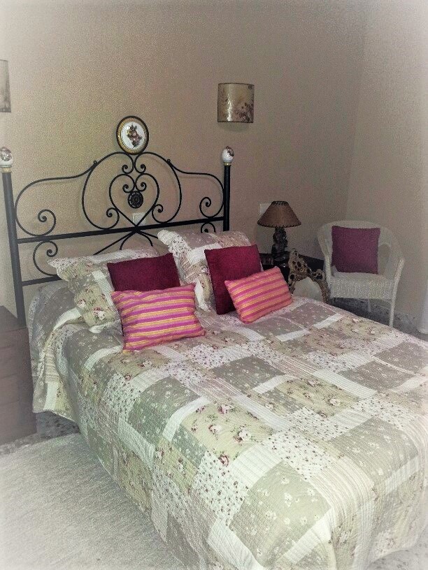 Bedroom featuring a double bed with a black wrought-iron headboard and patchwork bedding. A white wicker armchair and neutral-toned walls complete the space.