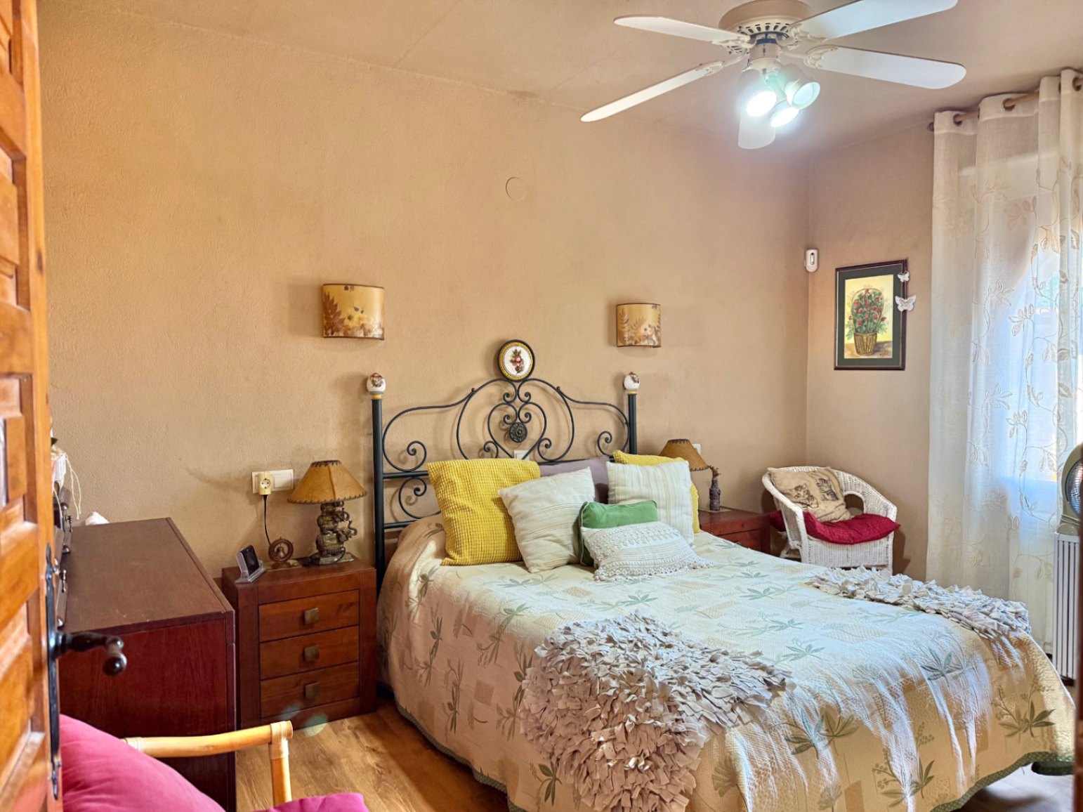 Bedroom featuring a wrought iron headboard, wooden bedside tables, and a ceiling fan. The room includes warm-toned walls, laminate flooring, and a wicker armchair.