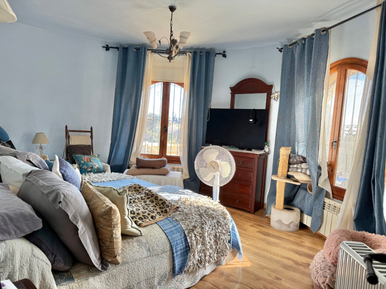 Bright bedroom featuring a curved wall with arched wooden windows, laminate flooring, and classic wooden furniture. Natural light fills the space through blue curtains.