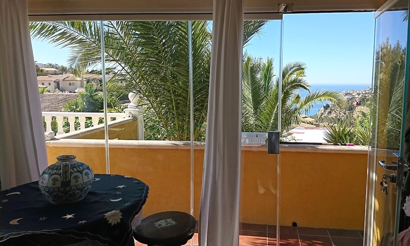 Enclosed terrace with frameless glass curtains, featuring an occasional table and panoramic views of the Mediterranean Sea and coastal hills.