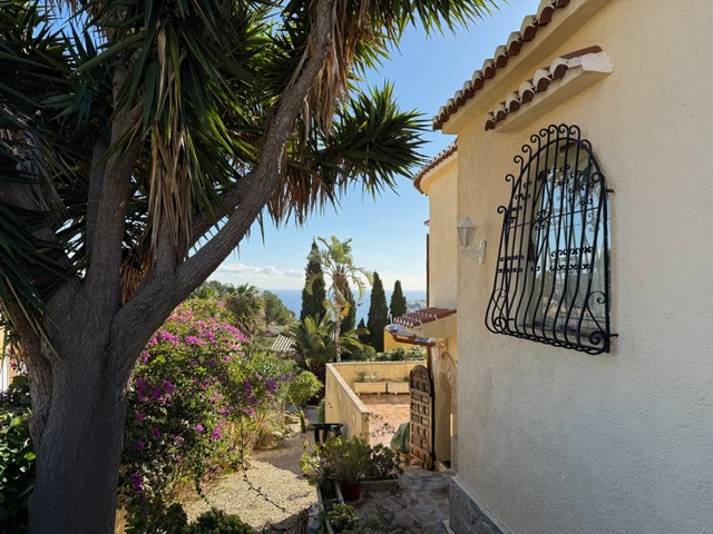 Exterior facade featuring traditional wrought iron window grilles, framed by mature Mediterranean gardens and distant sea views.