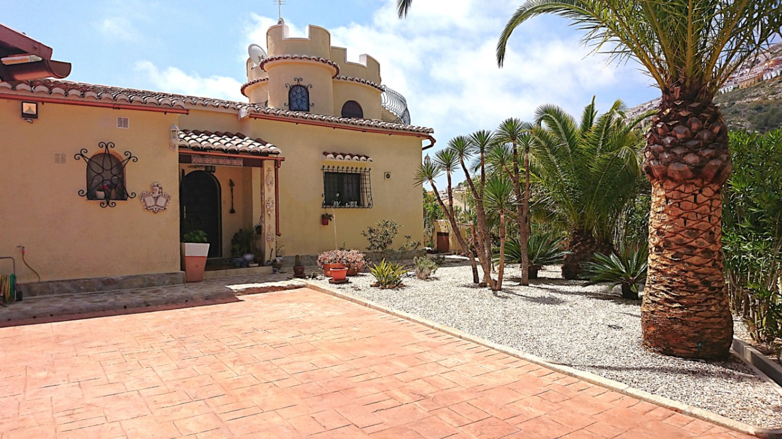 Exterior view featuring a crenellated tower, terracotta-paved entrance, and a Mediterranean gravel garden with mature palm trees.