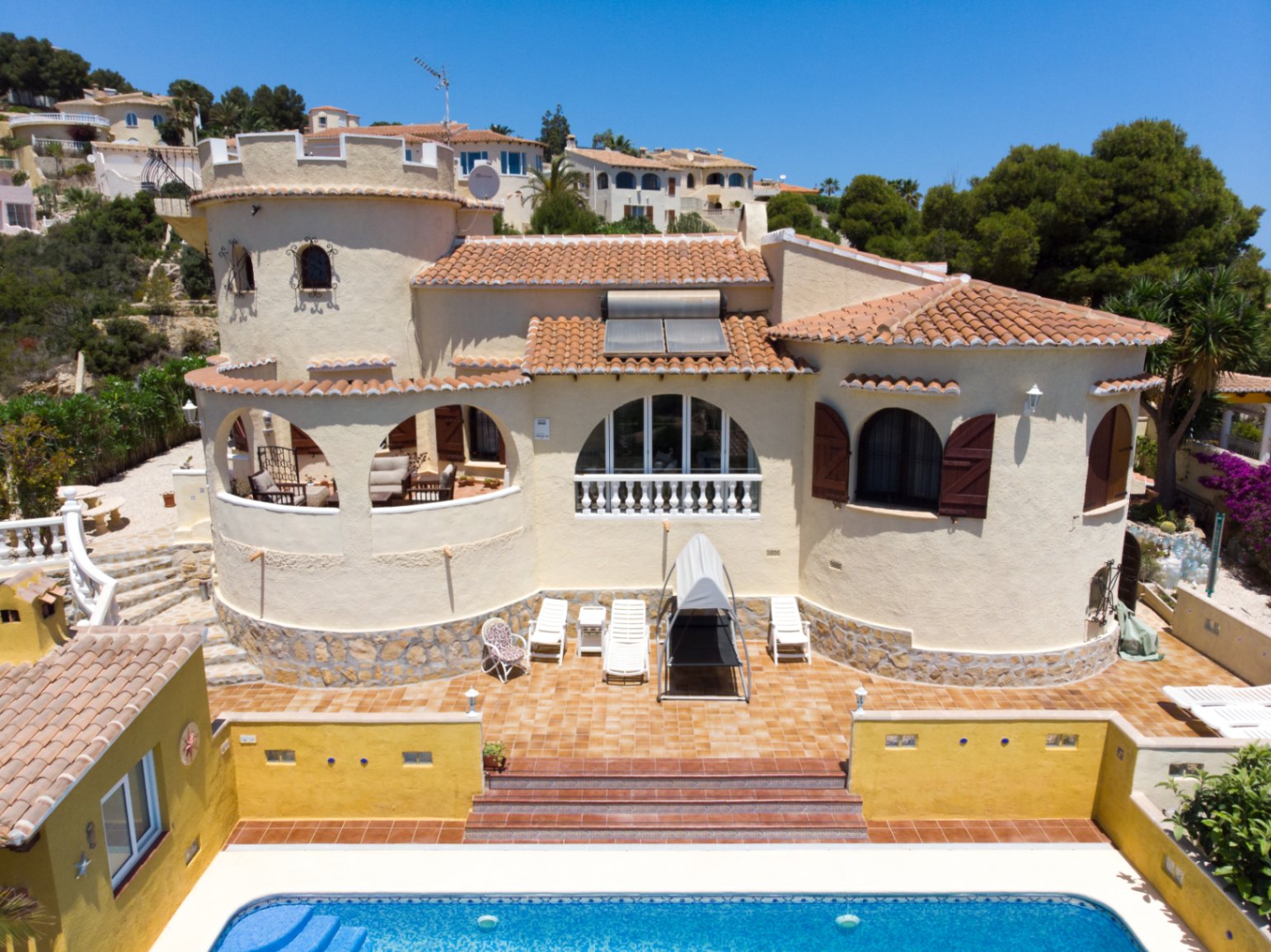 Exterior view of the Mediterranean villa featuring a distinctive tower, arched terraces, and a tiled pool. Includes terracotta roofing with solar panels and stone-clad walls.