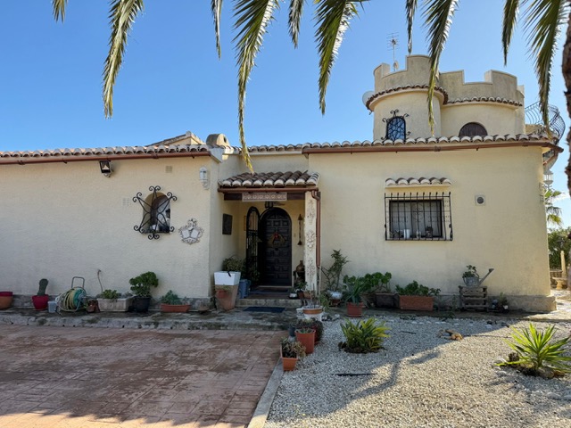 Exterior view of the villa featuring a traditional Mediterranean facade, a distinctive tower with battlements, and a wooden entrance door framed by potted plants.