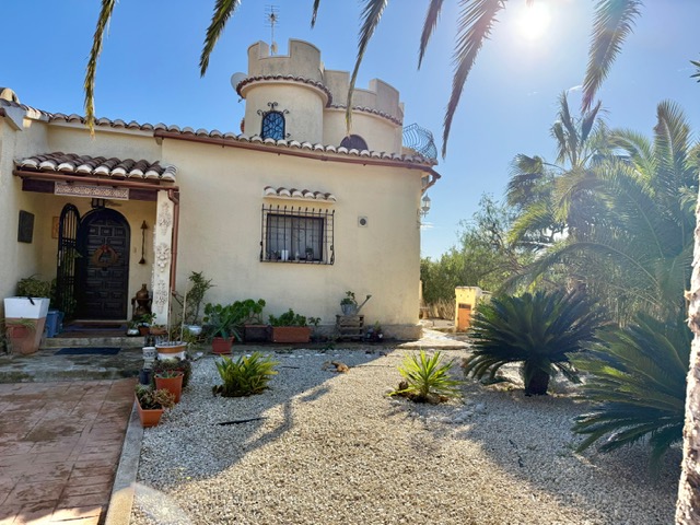 Main facade of the villa featuring a distinctive circular tower, tiled entrance porch, and a Mediterranean gravel garden with mature palm trees.