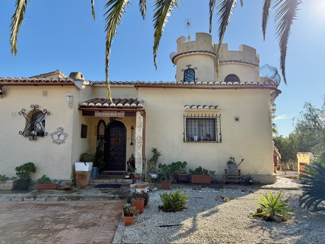Main facade of the villa featuring a distinctive crenellated tower, arched wooden entrance door, and traditional wrought iron window grilles set within a Mediterranean garden.