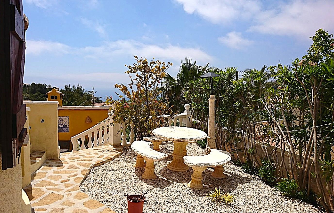Outdoor seating area featuring a carved stone table and benches set on gravel and natural stone paving, bordered by Mediterranean greenery and sea views.