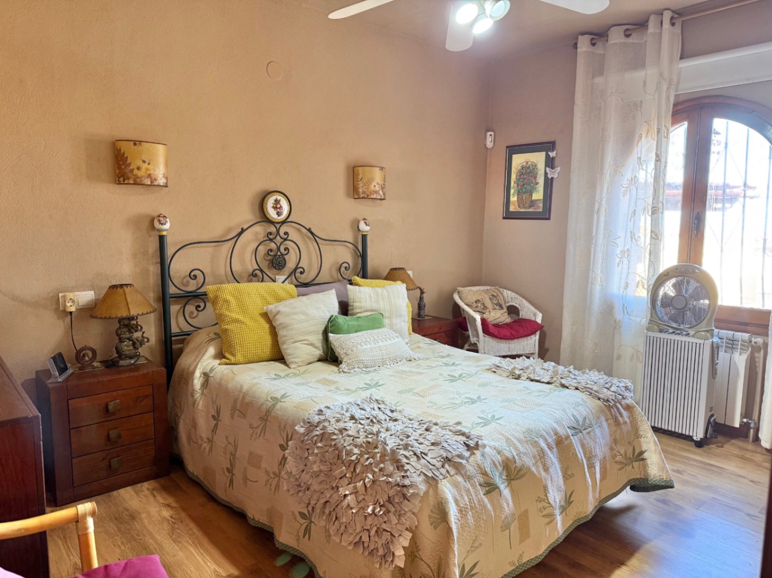 Rustic bedroom with a wrought iron headboard, wooden floors, and an arched window. Features a ceiling fan, radiator, and classic wooden nightstands.