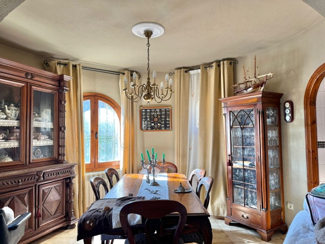 Rustic dining room featuring solid carved wood furniture, a glass display cabinet, and a central table illuminated by a brass chandelier and natural light.