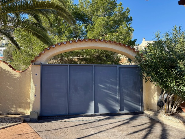 Sliding metal entrance gate set under a traditional Mediterranean arch with terracotta tiles, featuring a stamped concrete driveway and lush greenery.