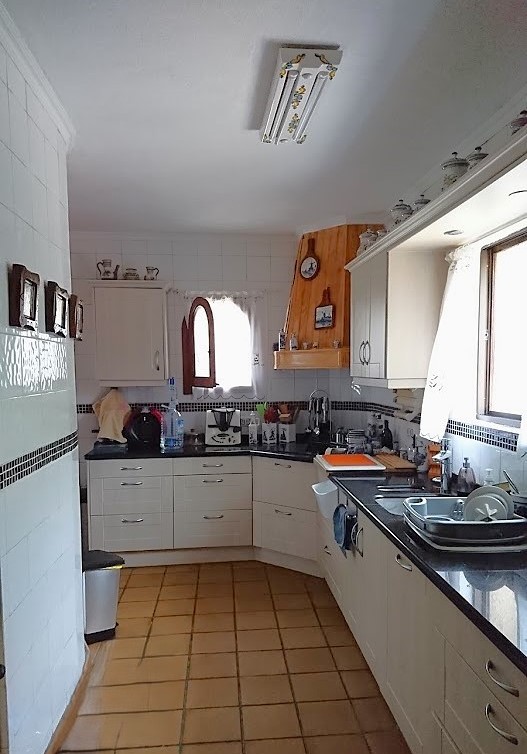 Traditional independent kitchen featuring white cabinetry, dark stone countertops, and terracotta floor tiles. Includes a wooden range hood cover and tiled walls with decorative borders.