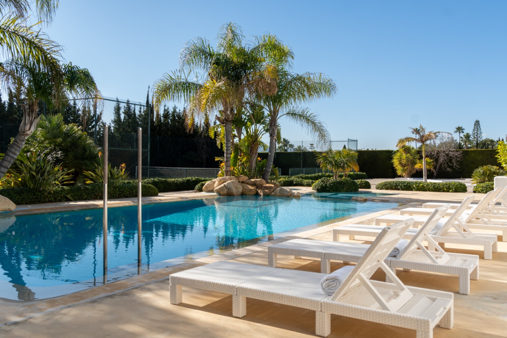 A 70sqm swimming pool area featuring white rattan sun loungers, tropical palm trees, and natural rock accents, with a private tennis court in the background.