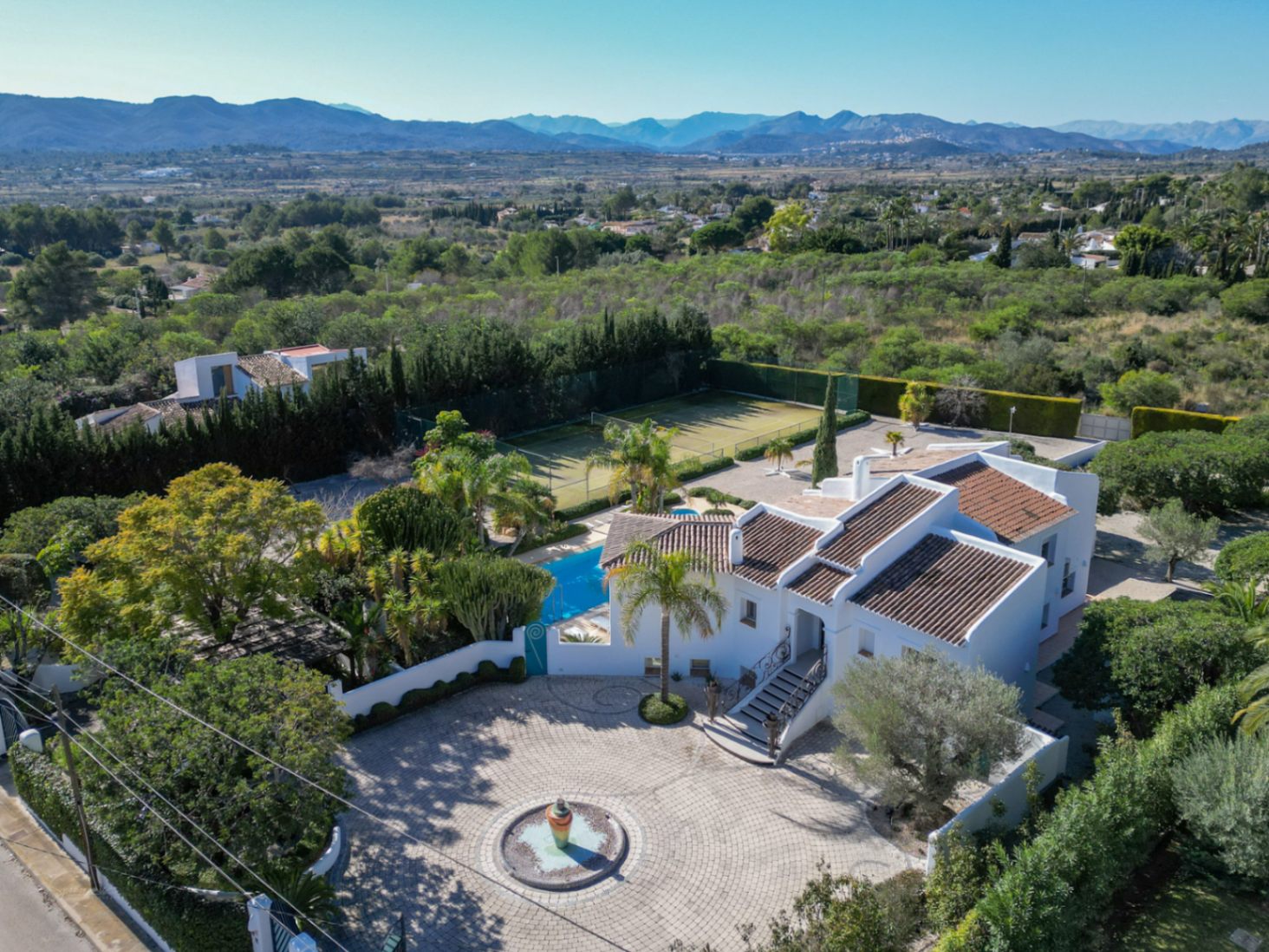 Aerial perspective of the Ibiza-style villa showing the paved entrance with a central fountain, private tennis court, and pool area overlooking the valley.