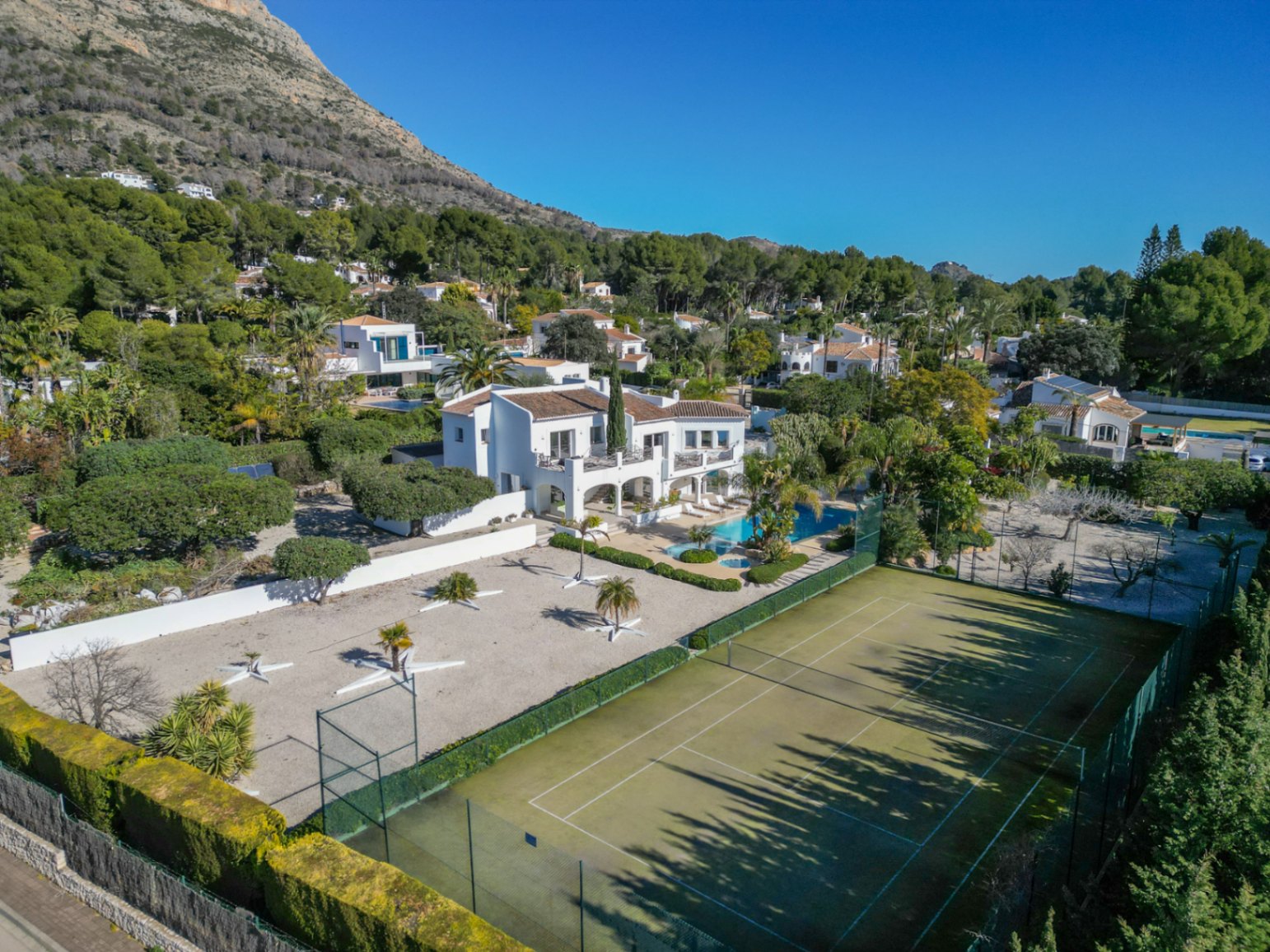 Aerial view of the Ibiza-style villa featuring a private tennis court, swimming pool with palm trees, and the Montgó mountain backdrop.