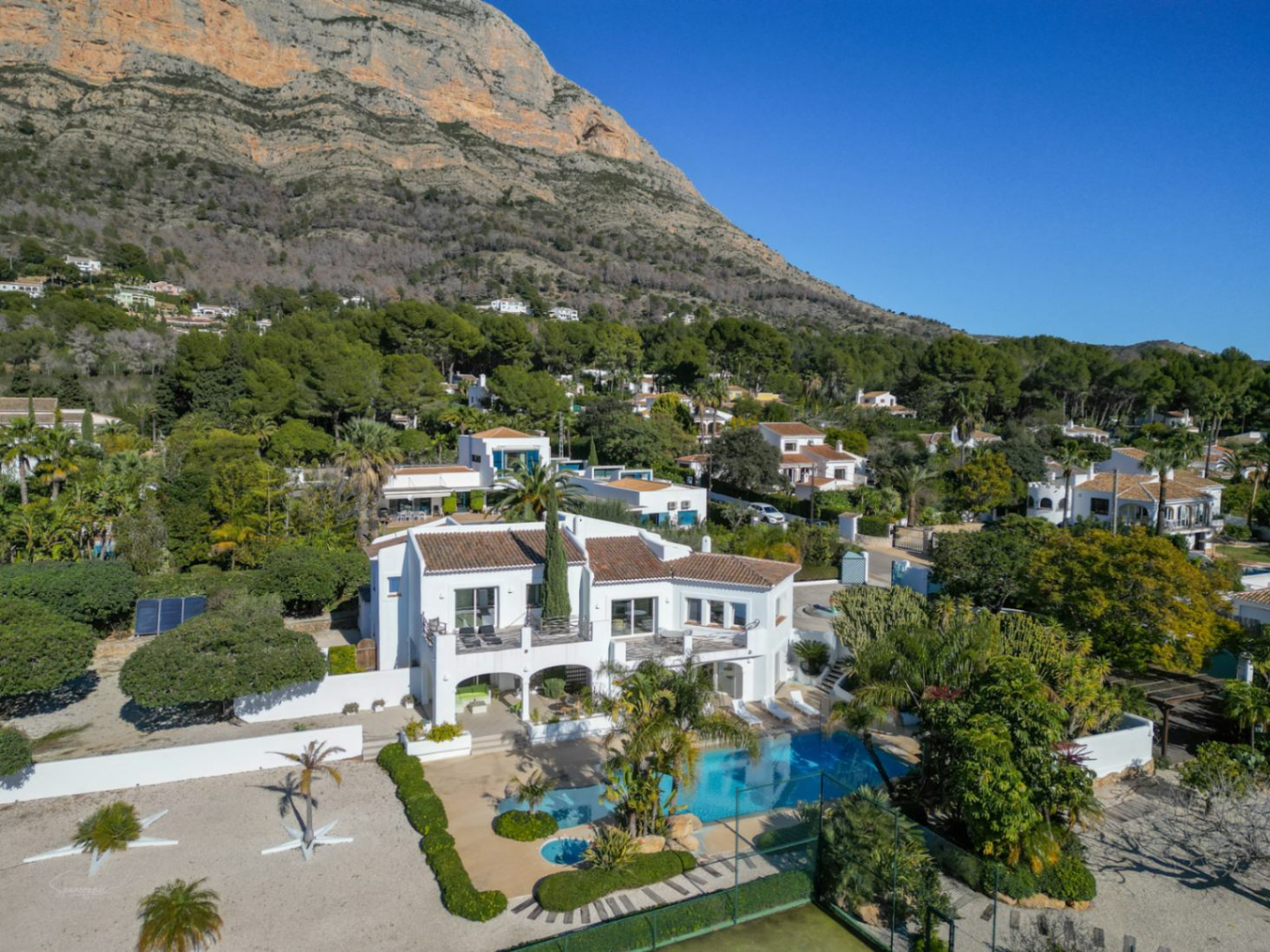 Aerial view of the Ibiza-style villa featuring white architecture, a private swimming pool, tennis court, and the majestic Montgó mountain as a backdrop.