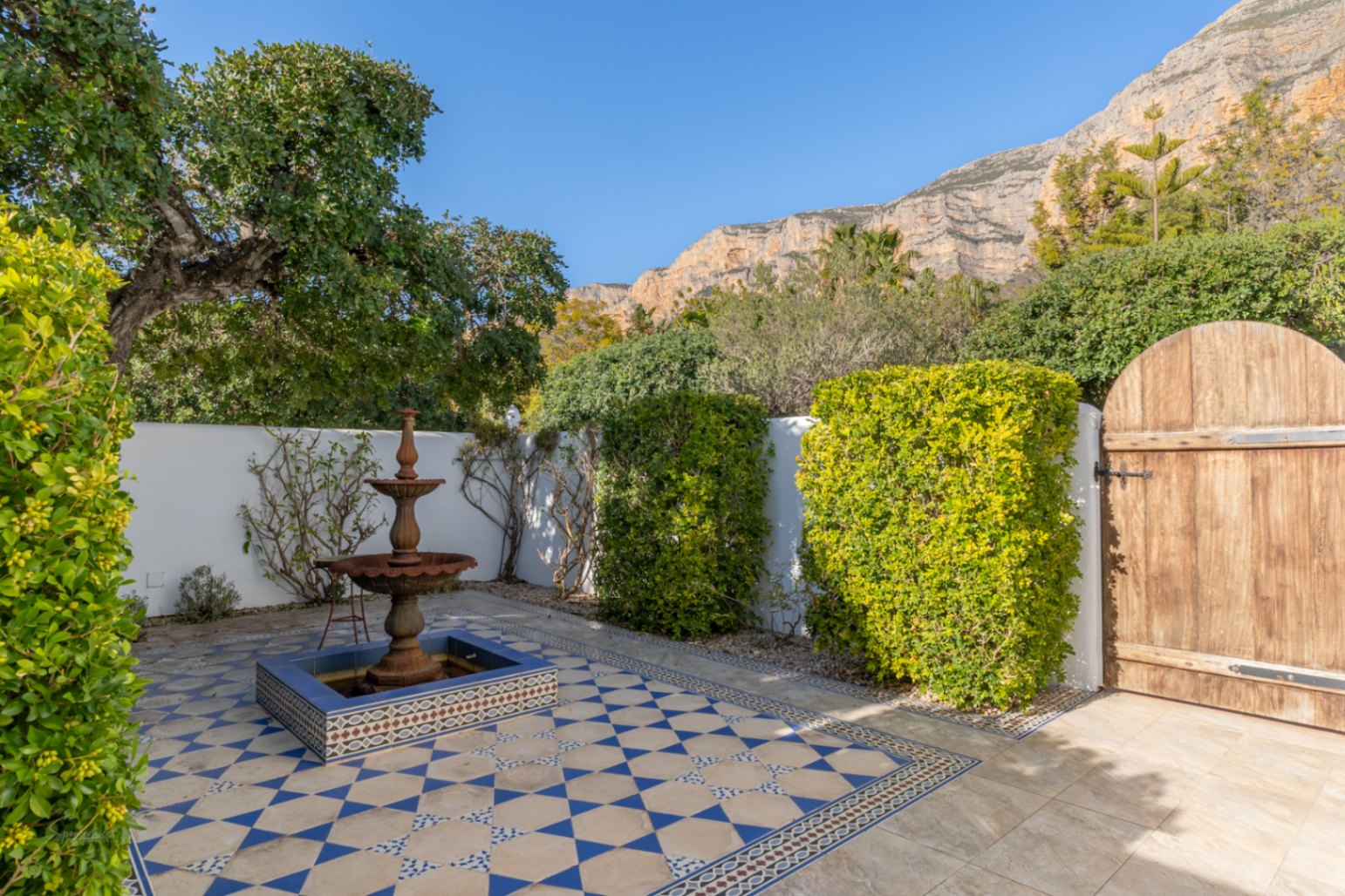 Andalusian-style courtyard featuring a three-tiered fountain, blue and white patterned tiling, and a rustic wooden gate set against the Montgó mountain.
