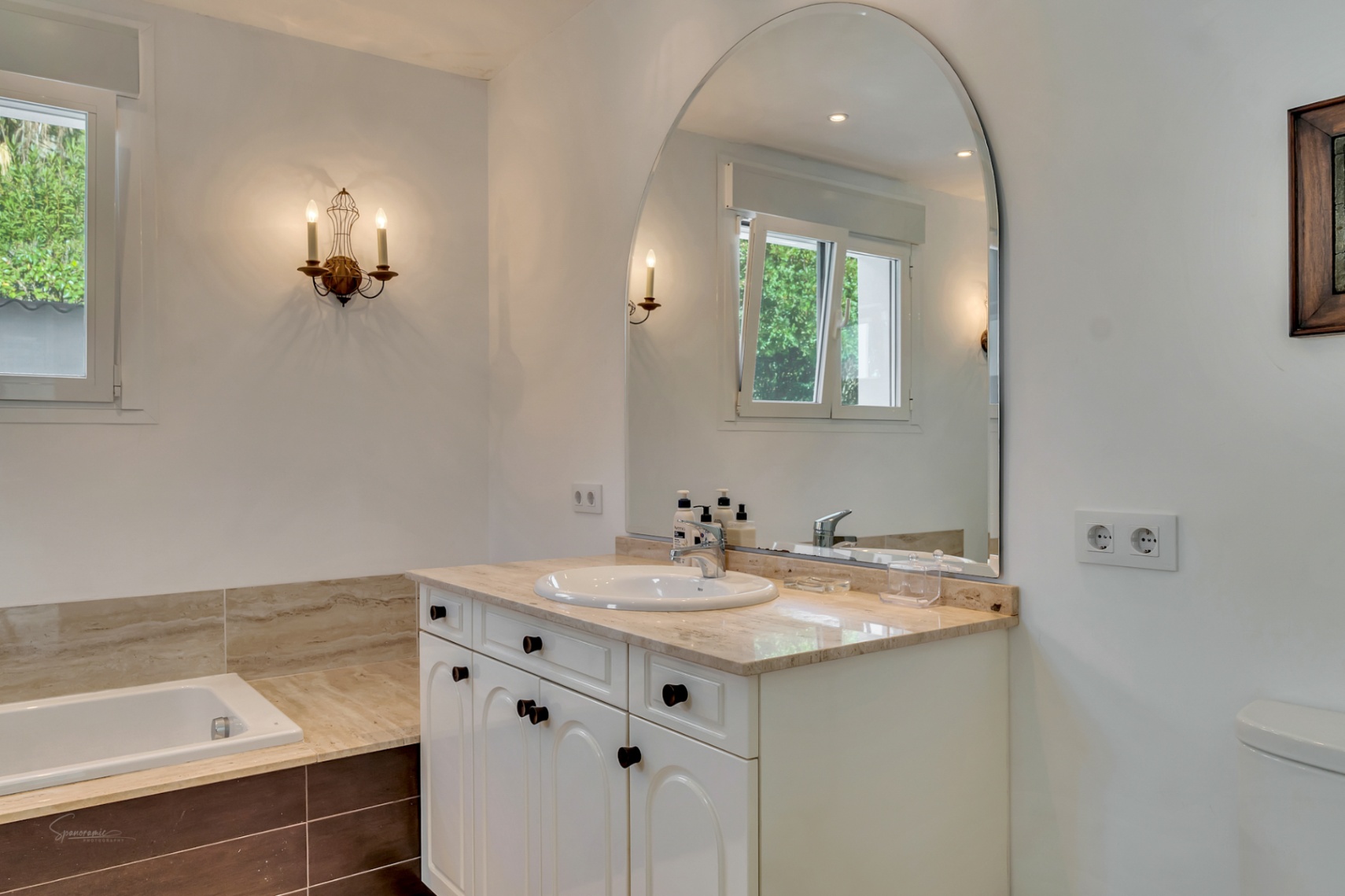 Bathroom featuring a marble vanity, large arched mirror, and classic wall sconces. The space includes an integrated bathtub and natural light from the side window.