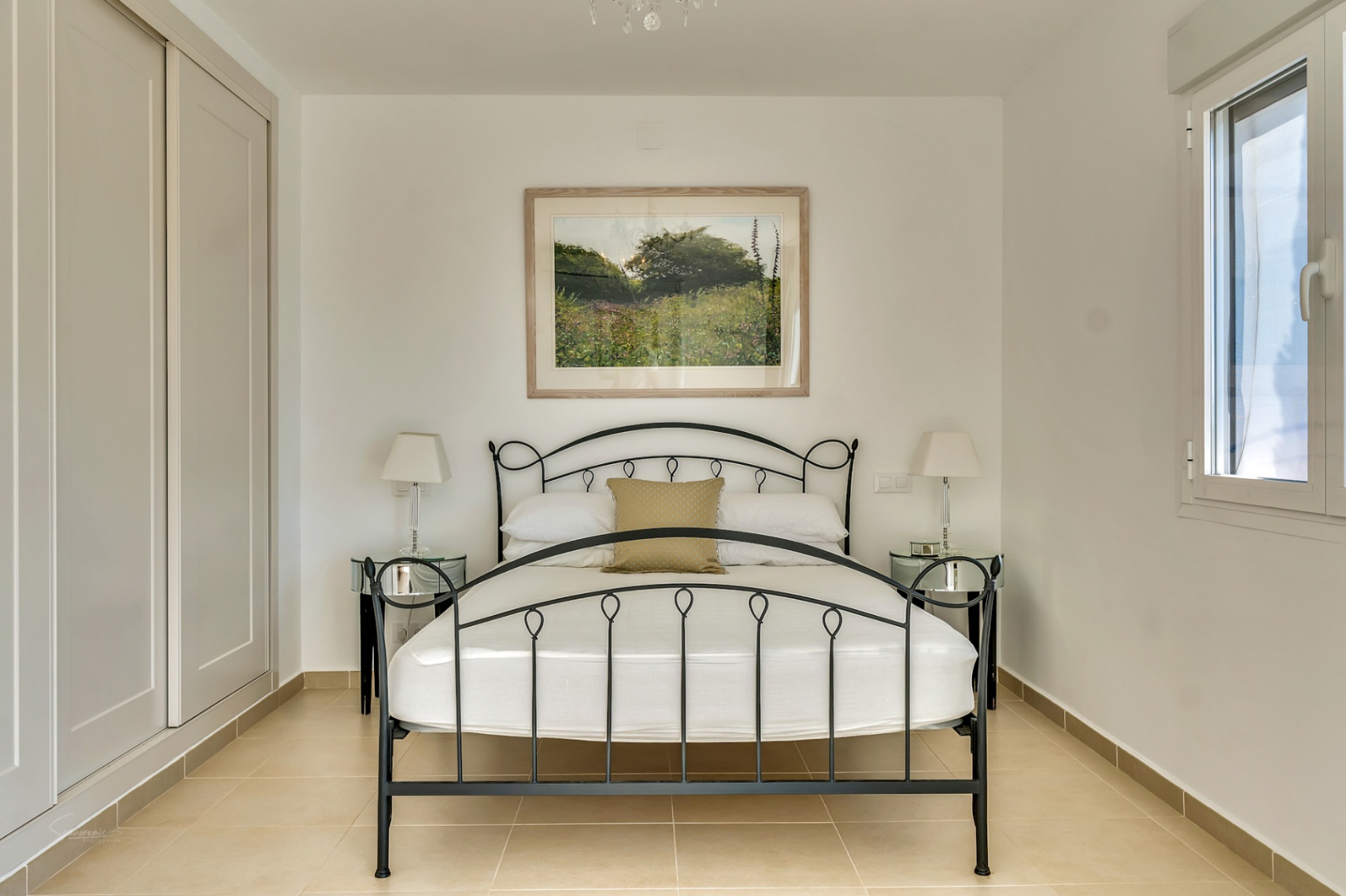 Bedroom featuring a black wrought iron bed frame, mirrored nightstands, and floor-to-ceiling built-in wardrobes. Neutral tiling and natural light define the space.