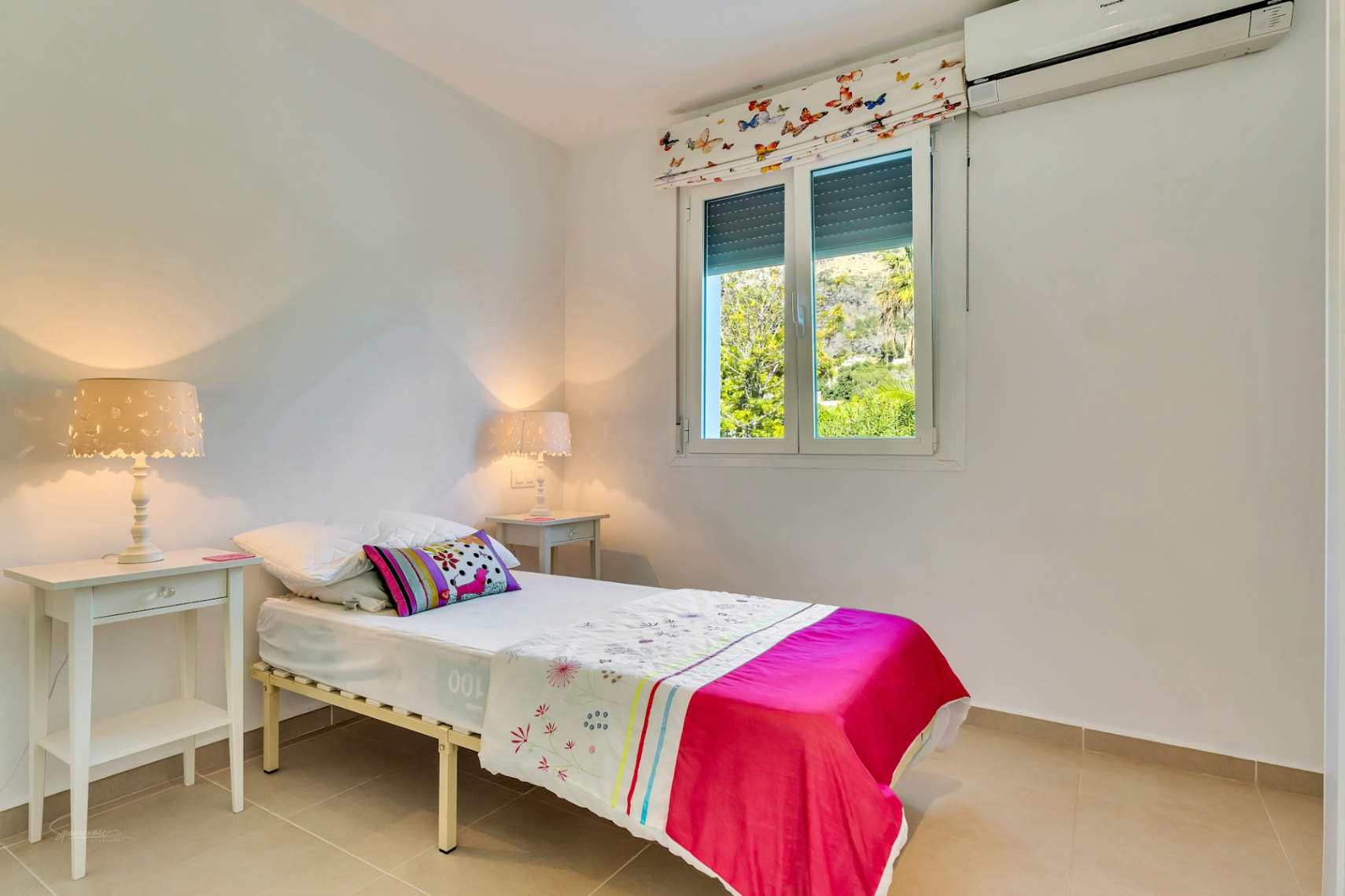 Bedroom featuring a single bed, split air conditioning, and a window with garden views. Includes white bedside tables and lamps with butterfly patterns.