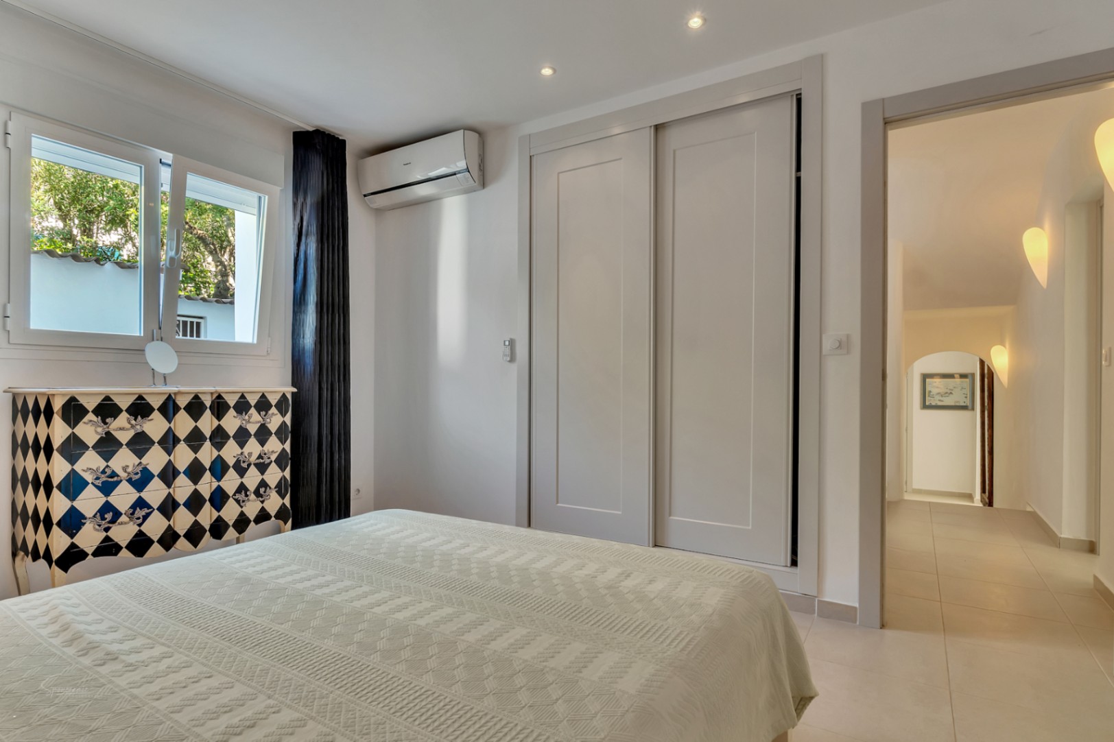 Bedroom featuring built-in sliding wardrobes, split air conditioning, and a distinctive harlequin dresser. The window provides natural light and garden views.