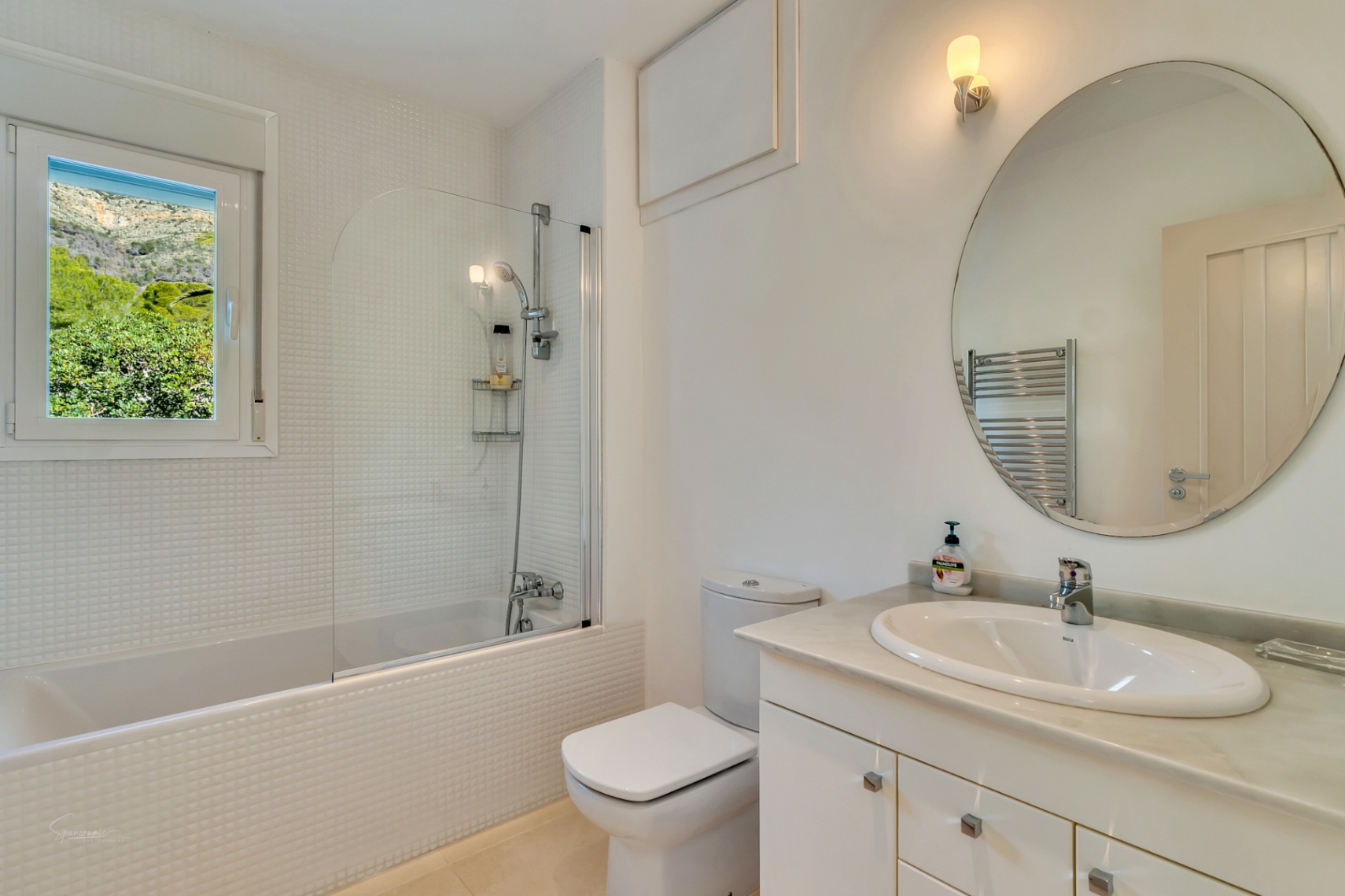 Bright bathroom featuring white mosaic tiling, a bathtub with glass partition, and a marble vanity. The window offers a direct view of the Montgó mountain.