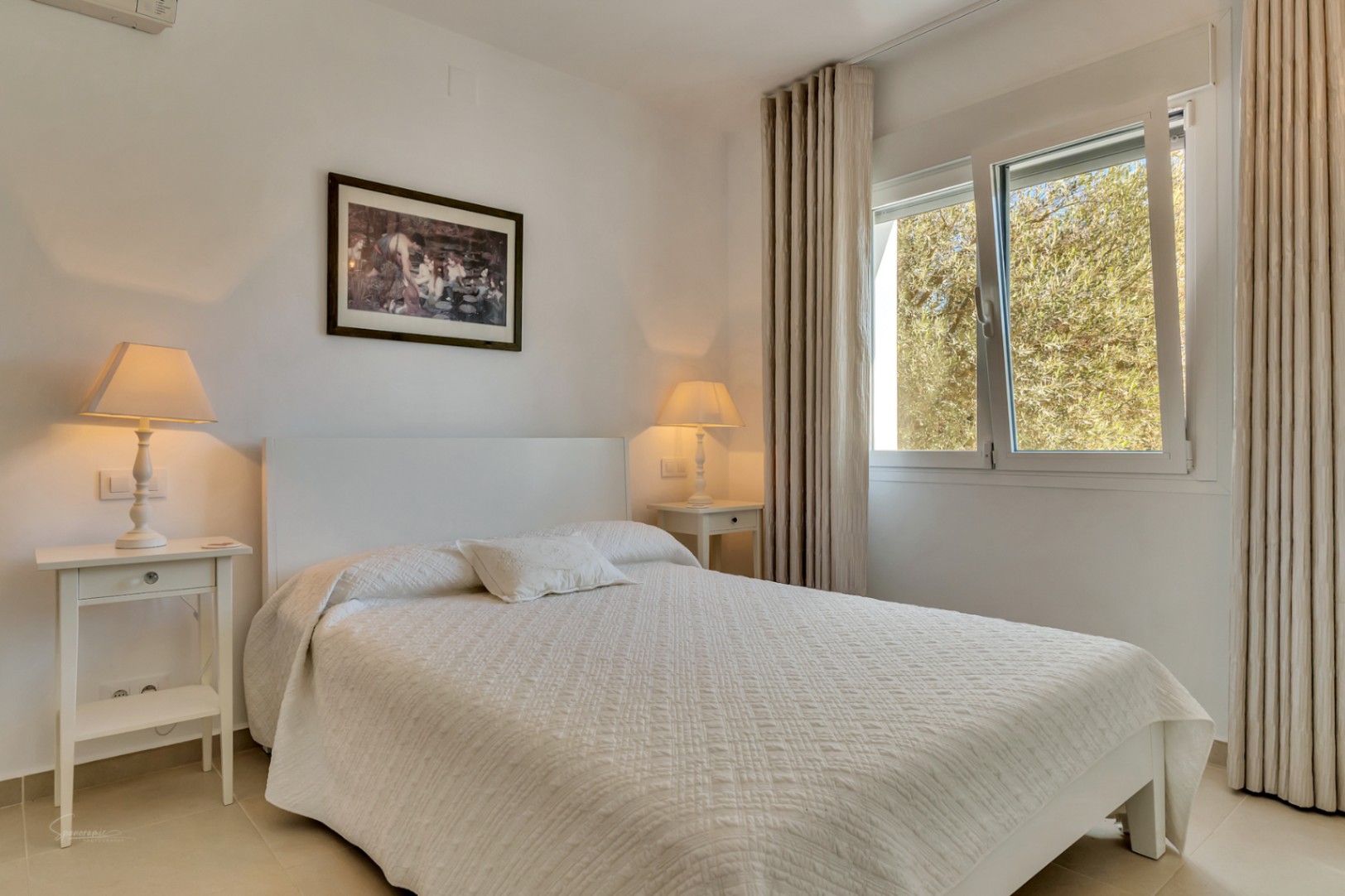 Bright bedroom featuring a white bed with textured linens, matching nightstands, and warm lighting. A large window provides natural light and views of the garden greenery.