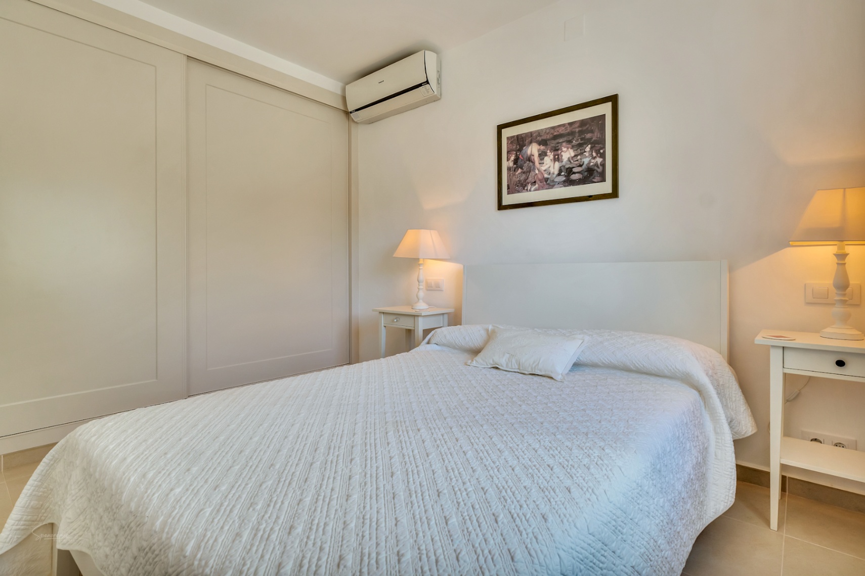 Bright bedroom featuring large built-in sliding wardrobes, split-system air conditioning, and elegant white bedside tables with warm ambient lighting.