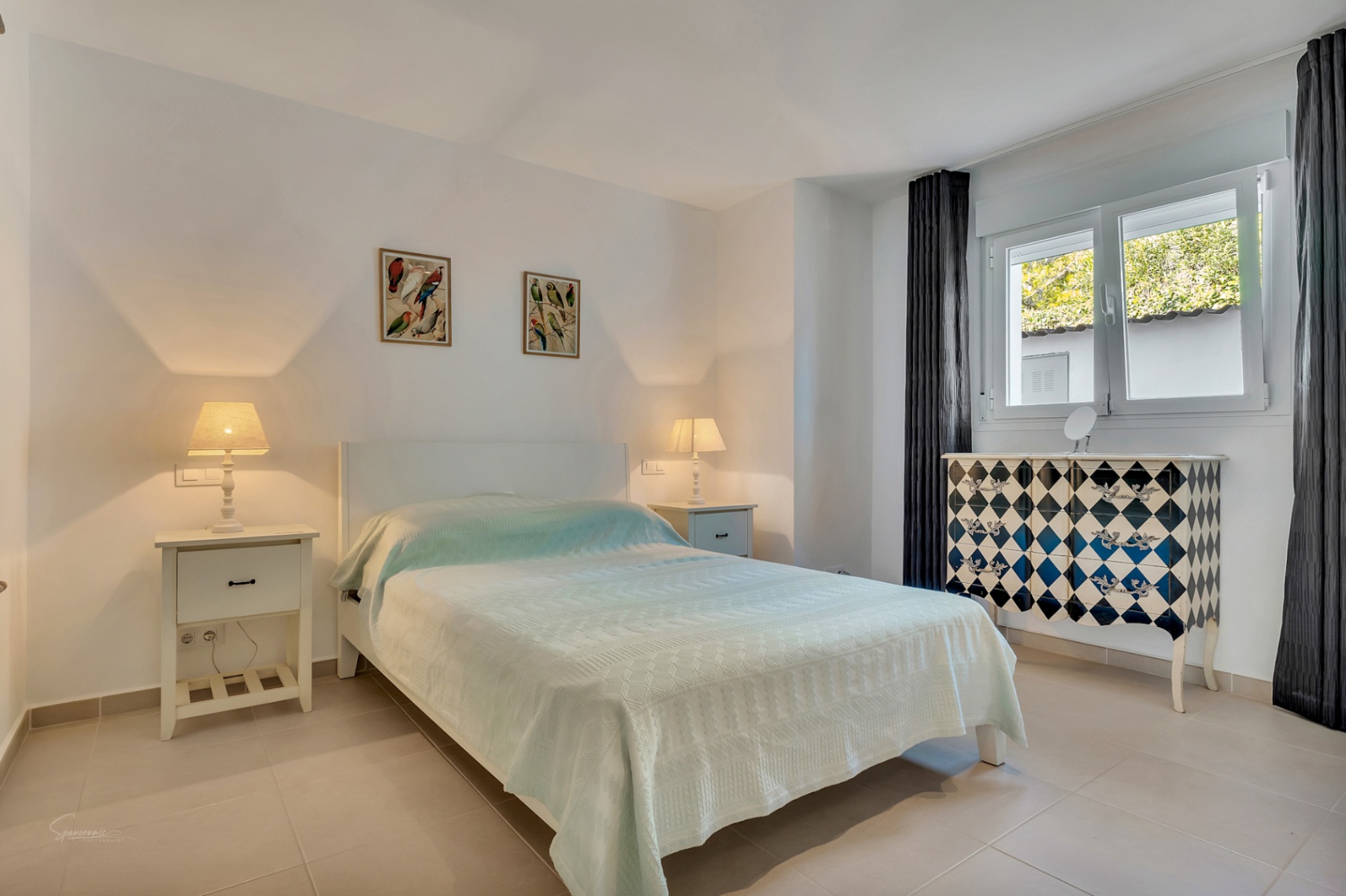Bright bedroom featuring tiled flooring, a double bed with white nightstands, and a distinctive harlequin-patterned chest of drawers by the window.