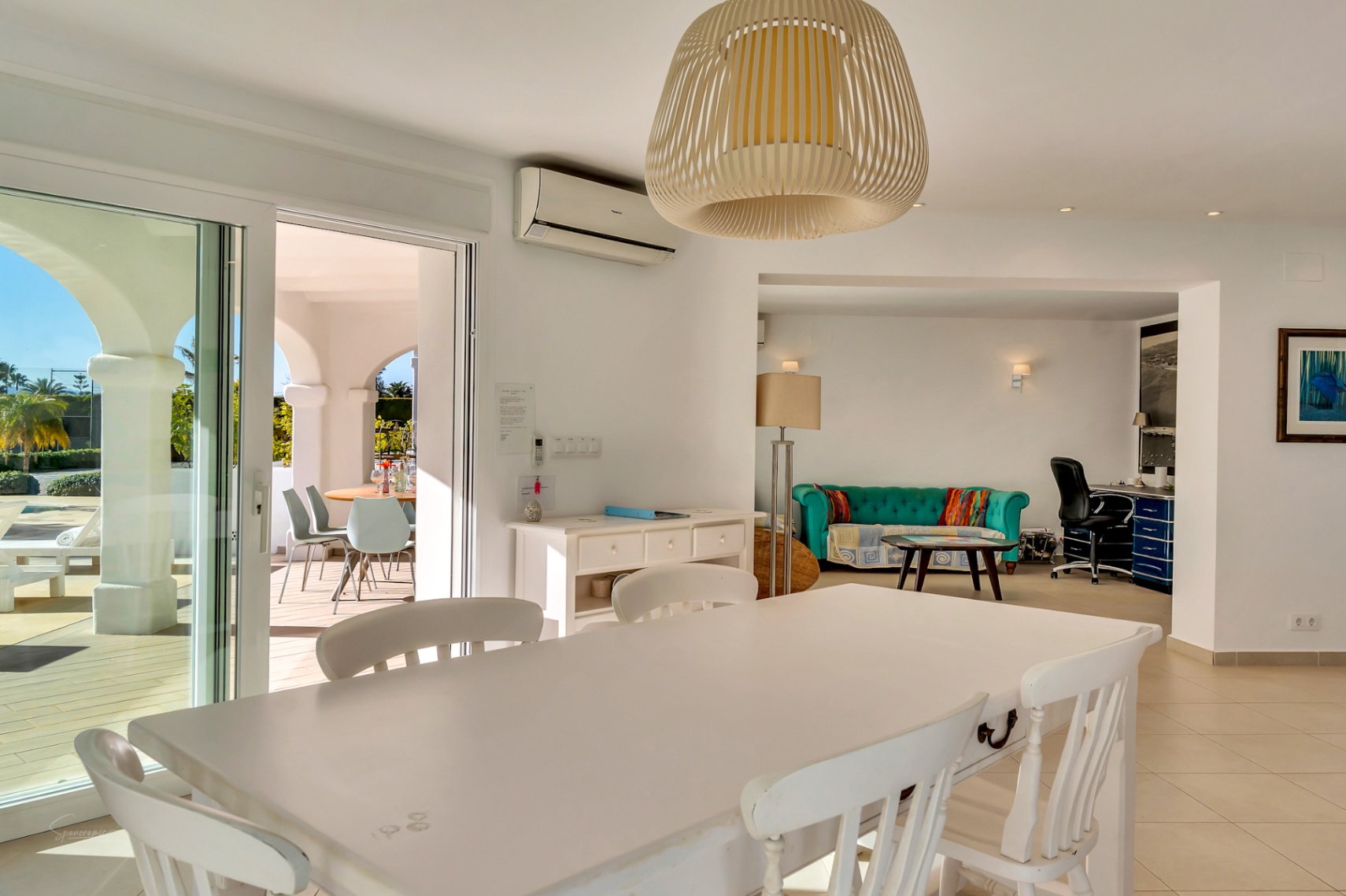 Bright dining area featuring a white wooden table and wicker pendant light, opening onto an arched naya and an integrated living space with a home office.