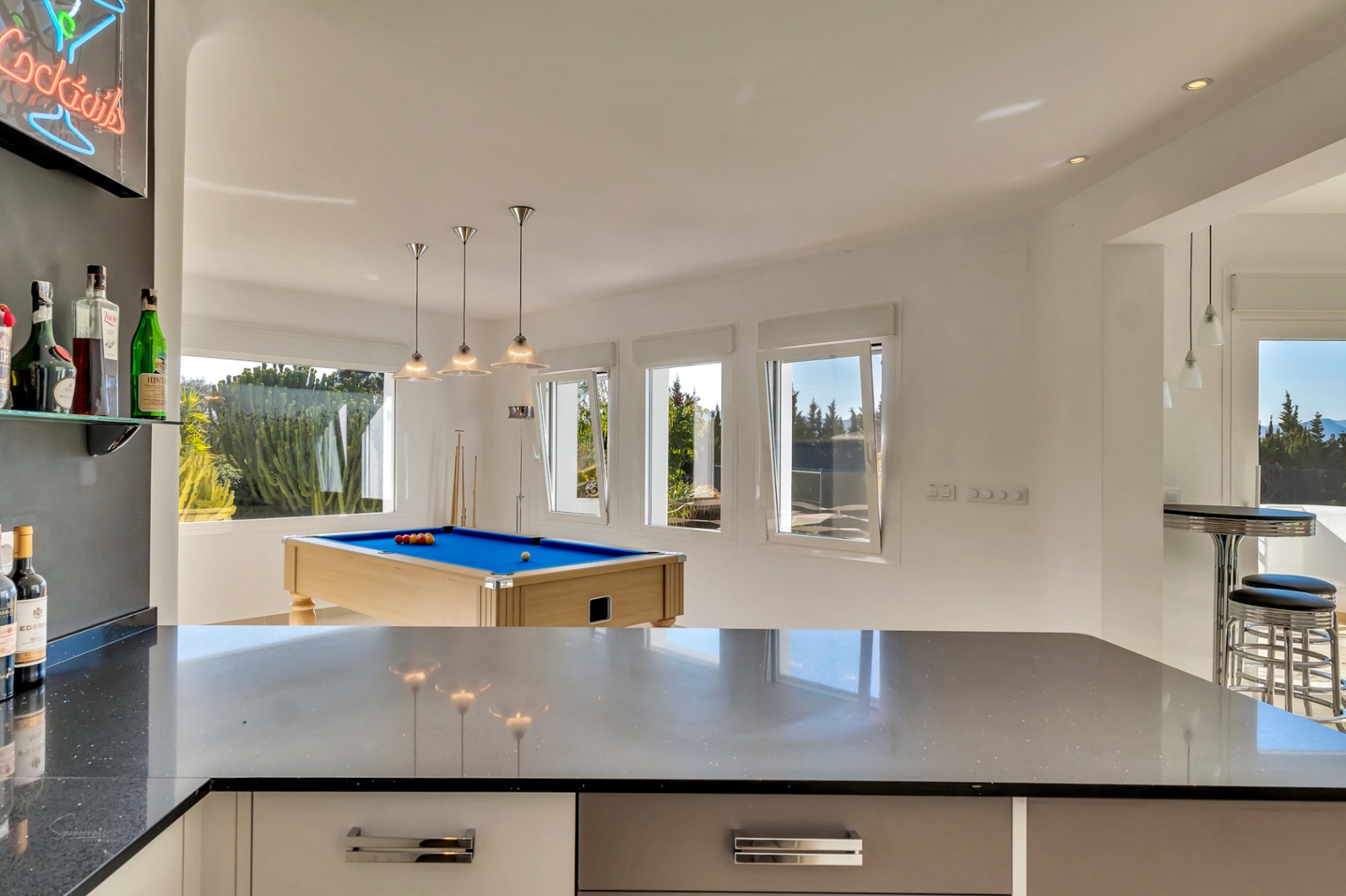 Bright entertainment space featuring a professional pool table, a sleek bar area with dark countertops, and large windows providing natural light.