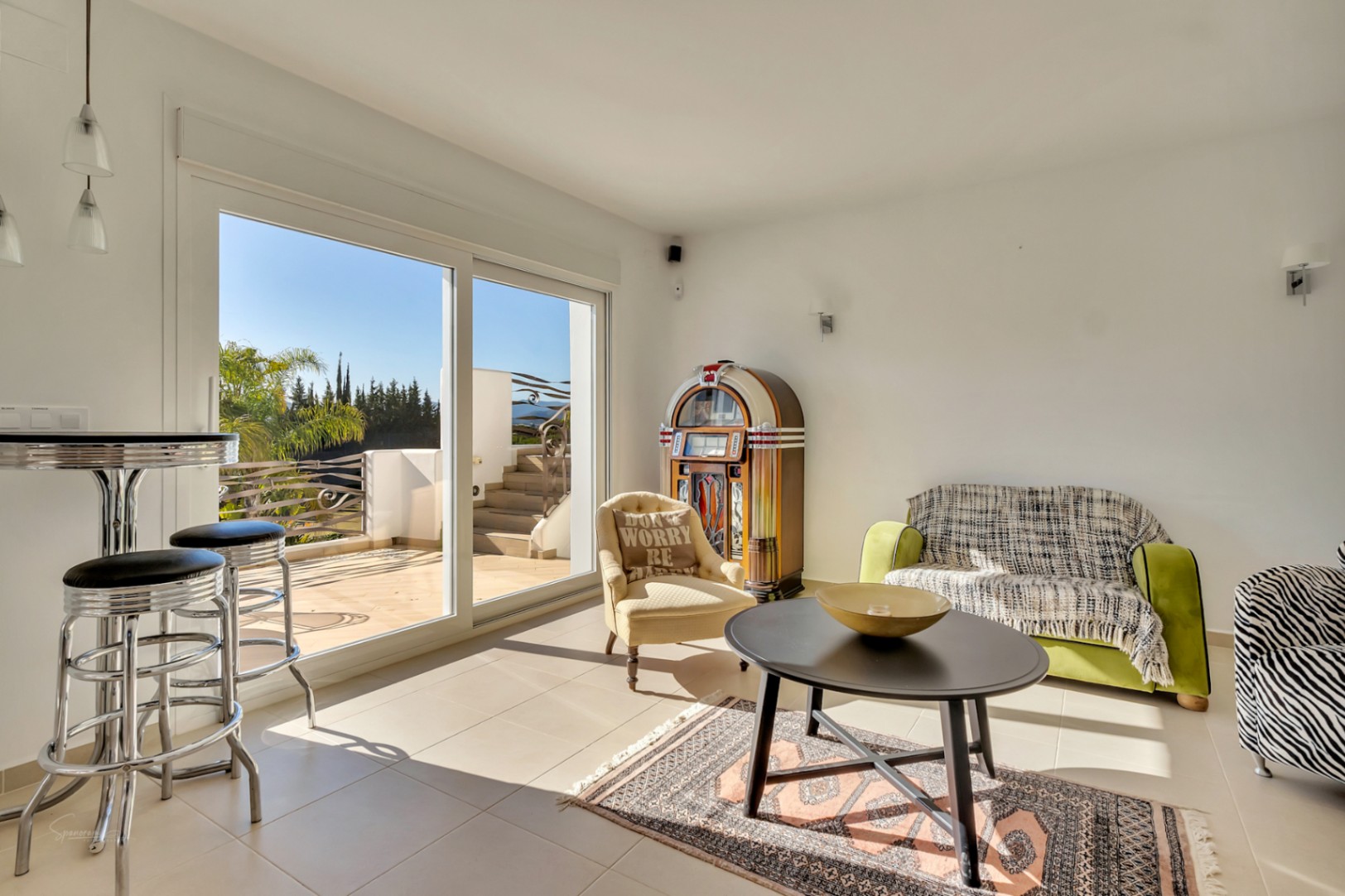 Bright living space featuring a vintage jukebox, eclectic seating, and large sliding glass doors that open onto a sunny terrace with garden views.