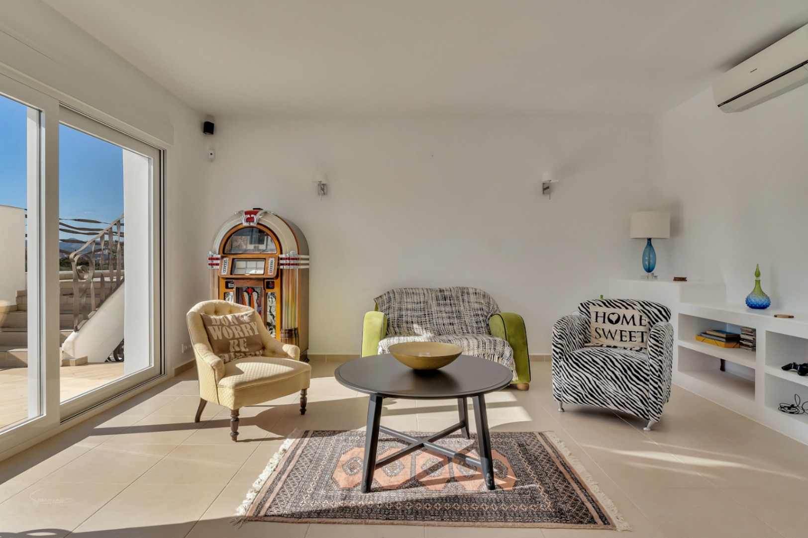 Bright lounge area featuring eclectic seating, a vintage jukebox, and direct terrace access. Includes integrated shelving and split-system air conditioning.