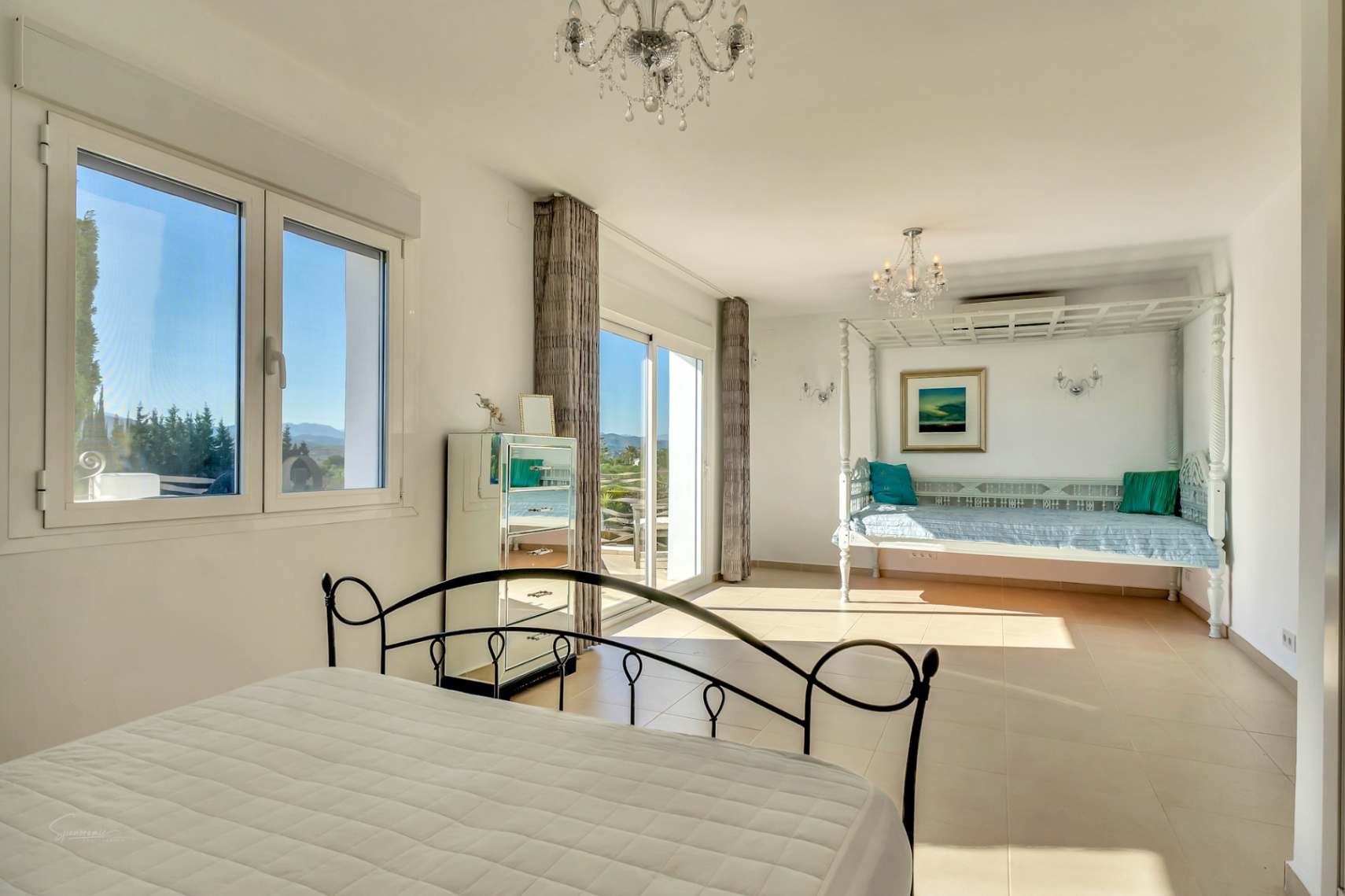 Bright master bedroom featuring a wrought iron bed, a white wooden daybed area, and sliding glass doors opening to a terrace with mountain views.