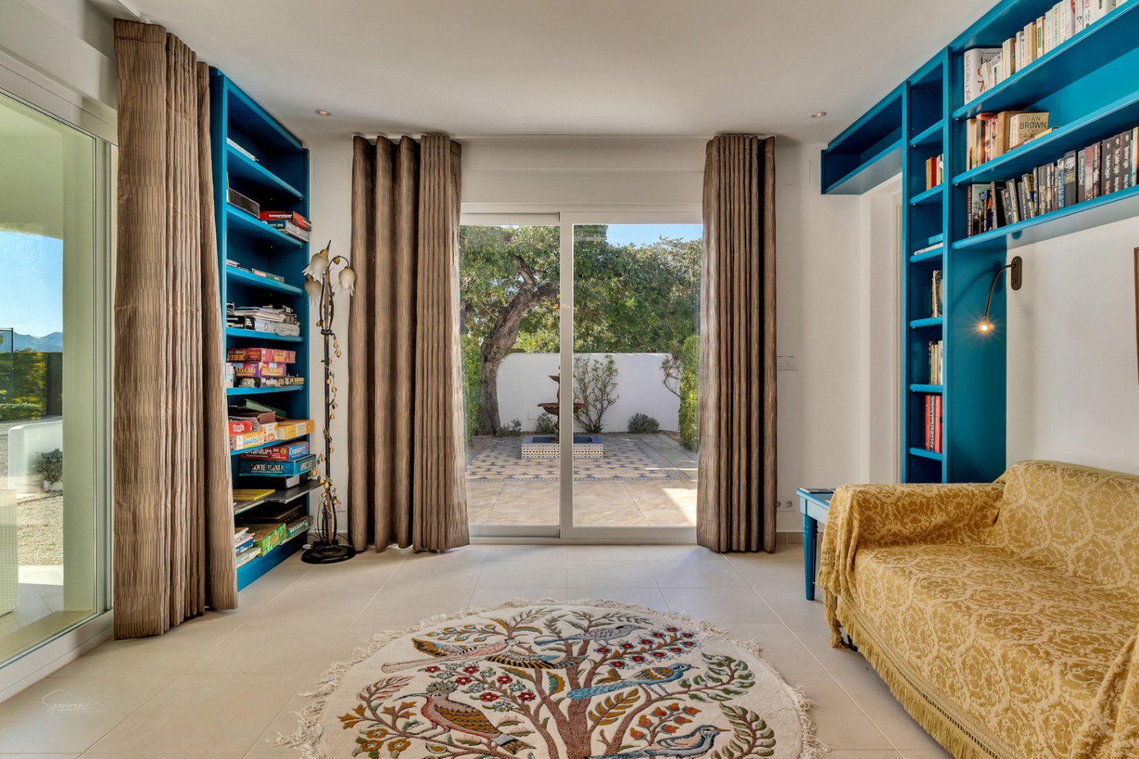 Bright study or lounge area featuring custom turquoise built-in shelving, a patterned sofa, and sliding doors opening onto a private courtyard with a fountain.