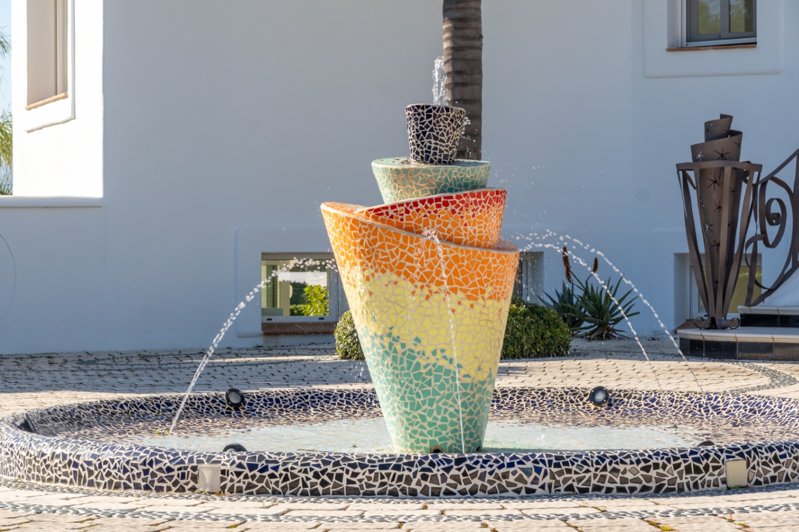 Contemporary tiered fountain featuring vibrant multi-colored mosaic tiles, set within a paved courtyard against the villa's white exterior.