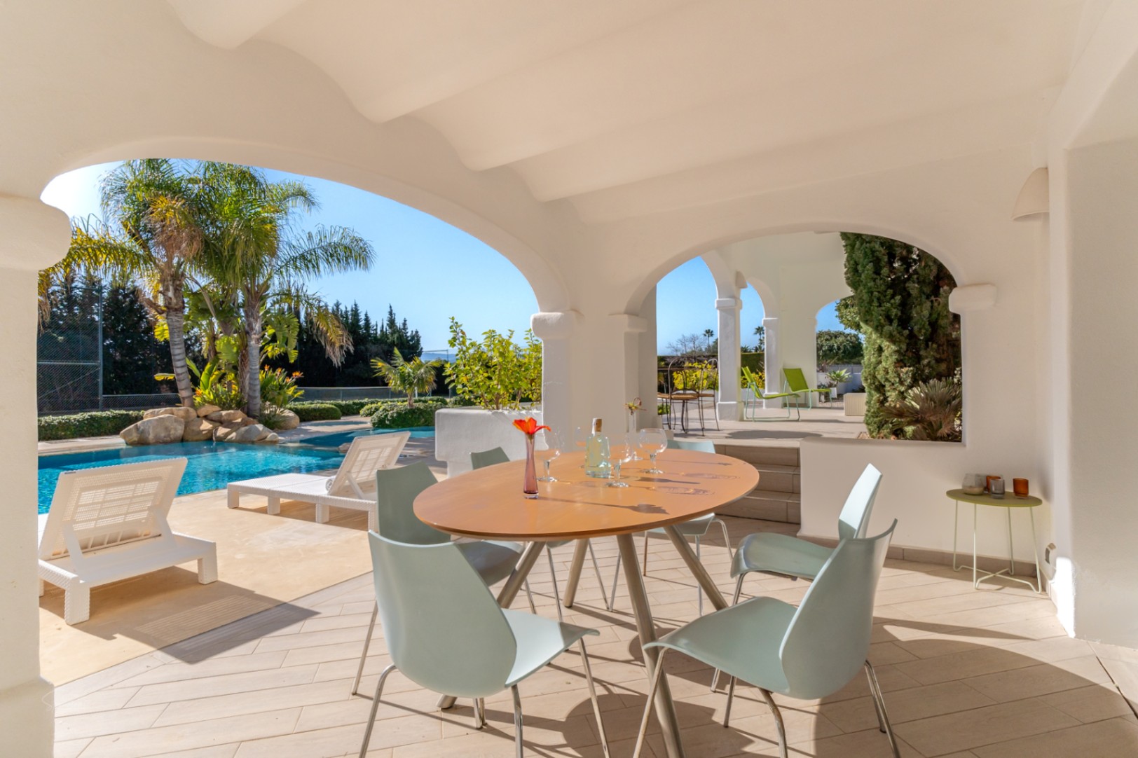 Covered naya featuring white arches and an outdoor dining area, overlooking the 70sqm swimming pool, sun loungers, and private tennis court.