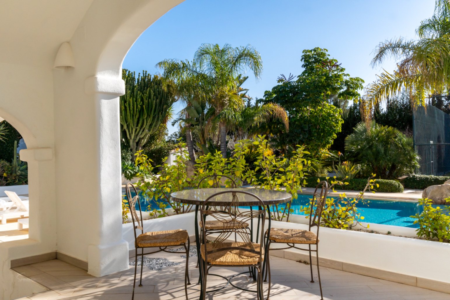 Covered naya featuring white arches and wrought-iron dining furniture, overlooking the private swimming pool and a lush garden with palm trees.