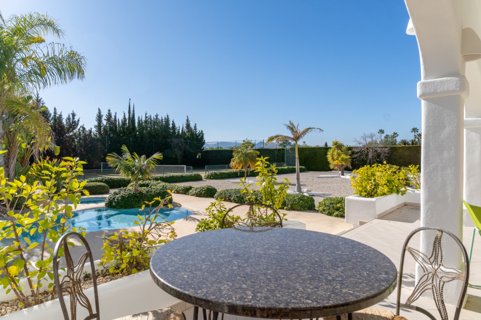 Covered terrace featuring a stone table and views overlooking the swimming pool, landscaped Mediterranean garden, and private tennis court.