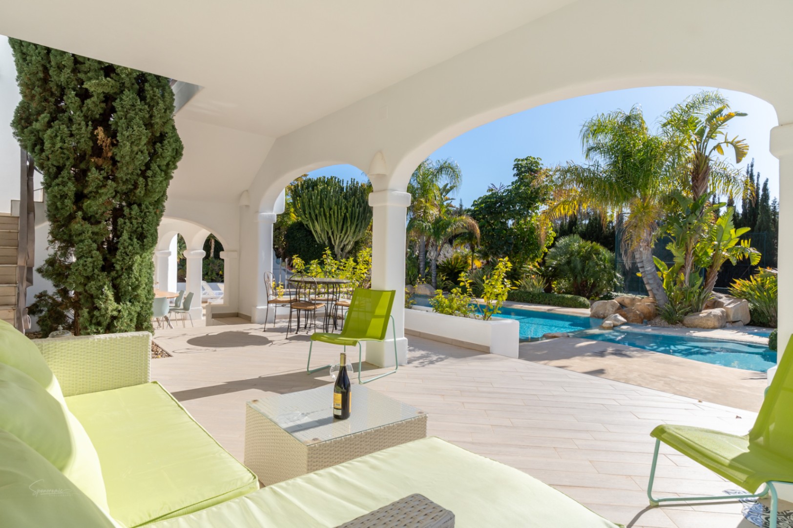Covered terrace with white arches and tiled flooring, featuring lime green lounge seating overlooking the swimming pool and lush tropical garden.