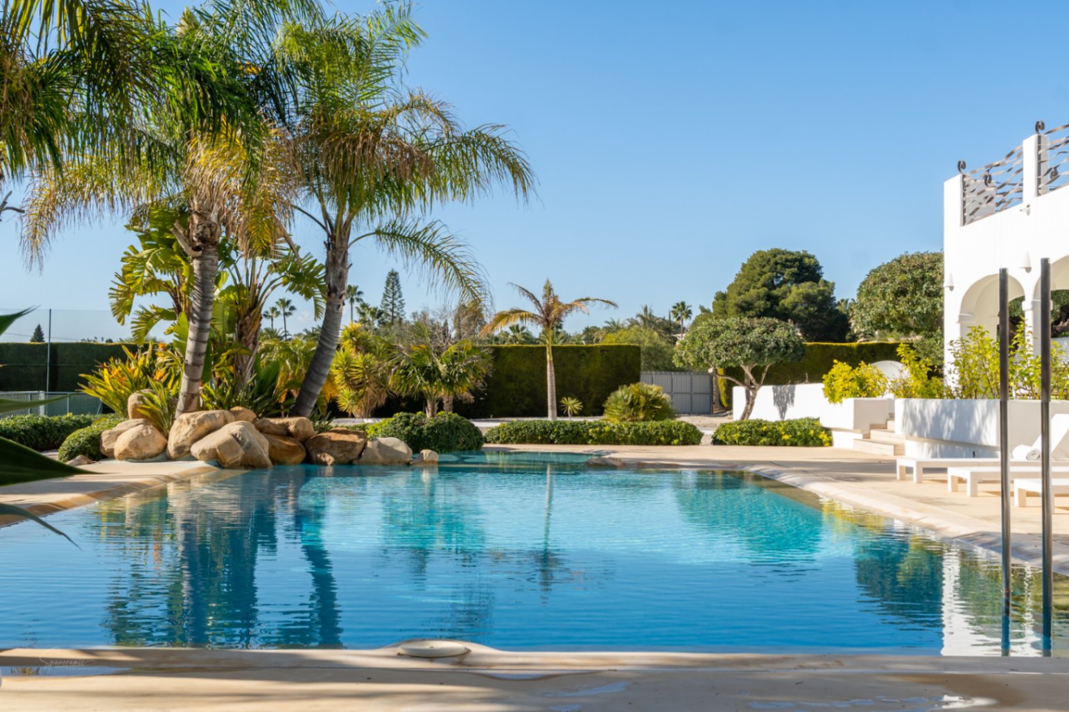 Designer swimming pool with beach entry, surrounded by tropical palms, rock features, and a manicured garden adjacent to the white arched naya and tennis court.